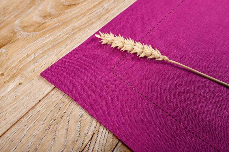 May include: A close-up of a purple linen placemat with a single stalk of wheat resting on top.