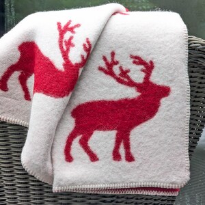 May include: White throw blanket with a red reindeer design. The blanket is folded and draped over a wicker chair.