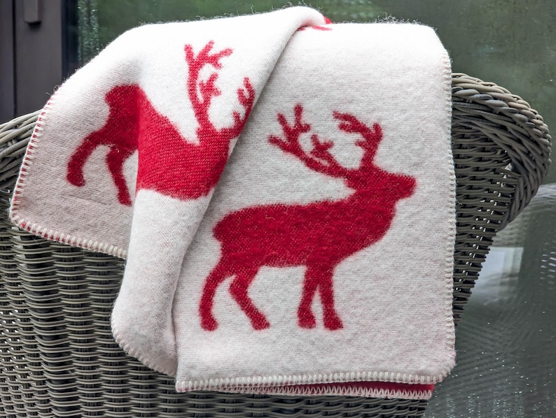 May include: White throw blanket with a red reindeer design. The blanket is folded and draped over a wicker chair.