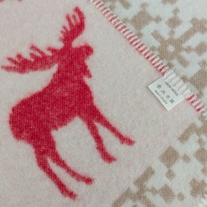 May include: A white throw blanket with a red moose design and a brown snowflake pattern. The blanket has a label that reads "100% Wool" and "Made in Lithuania".