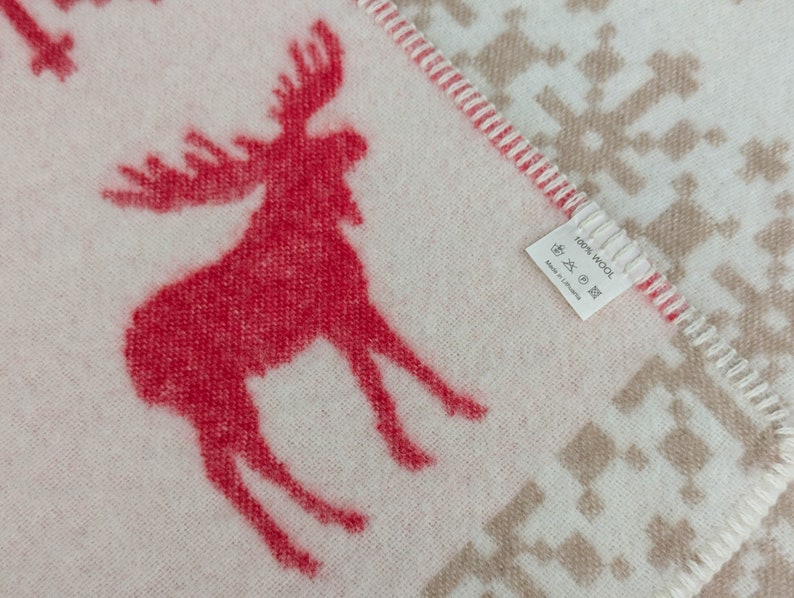 May include: A white throw blanket with a red moose design and a brown snowflake pattern. The blanket has a label that reads "100% Wool" and "Made in Lithuania".
