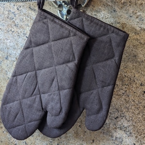 K&ouml;nnte beinhalten: Ein Paar braune, gesteppte Ofenhandschuhe, die an einem Haken h&auml;ngen. Die Handschuhe sind aus einem dicken, strapazierf&auml;higen Stoff gefertigt und haben ein gestepptes Design f&uuml;r zus&auml;tzlichen Schutz.