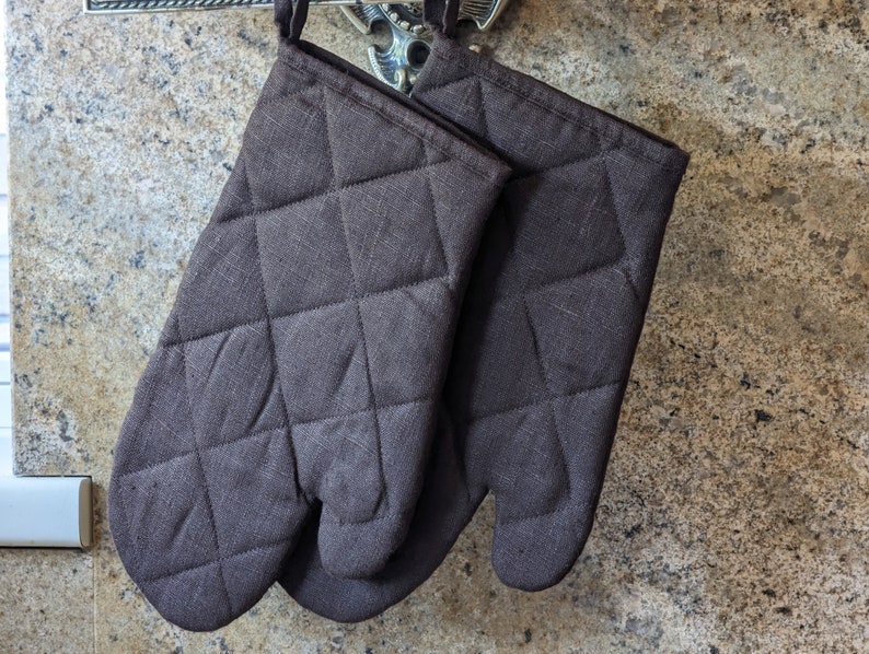 K&ouml;nnte beinhalten: Ein Paar braune, gesteppte Ofenhandschuhe, die an einem Haken h&auml;ngen. Die Handschuhe sind aus einem dicken, strapazierf&auml;higen Stoff gefertigt und haben ein gestepptes Design f&uuml;r zus&auml;tzlichen Schutz.