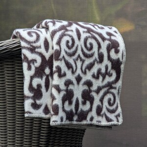 Puede incluir: Una manta de tiro blanca y marr&oacute;n con un dise&ntilde;o de damasco. La manta est&aacute; doblada y drapeada sobre una silla de mimbre.