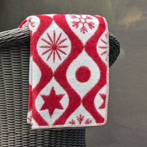 Puede incluir: Una manta plegada con estampado rojo y blanco sobre una silla de mimbre. La manta presenta un dise&ntilde;o repetitivo de formas geom&eacute;tricas rojas y patrones de copos de nieve, estrellas y rayos de sol sobre un fondo blanco. Los bordes de la manta est&aacute;n acabados con costuras blancas.