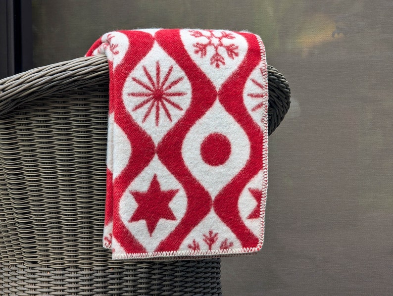 Puede incluir: Una manta plegada con estampado rojo y blanco sobre una silla de mimbre. La manta presenta un dise&ntilde;o repetitivo de formas geom&eacute;tricas rojas y patrones de copos de nieve, estrellas y rayos de sol sobre un fondo blanco. Los bordes de la manta est&aacute;n acabados con costuras blancas.