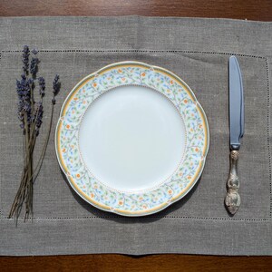 Könnte beinhalten: Ein weißer Porzellanteller mit einem floralen Muster in Gelb-, Blau- und Grüntönen. Der Teller liegt auf einem grauen Leinen-Platzset mit einem silbernen Messer und einem Lavendelstrauß.