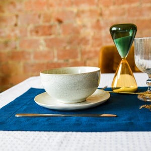 Puede incluir: Un cuenco de cer&aacute;mica blanca con un acabado moteado se encuentra sobre un mantel individual azul con un tenedor plateado y un reloj de arena dorado en el fondo. El mantel individual est&aacute; sobre un mantel blanco con un patr&oacute;n de lunares blancos y negros.