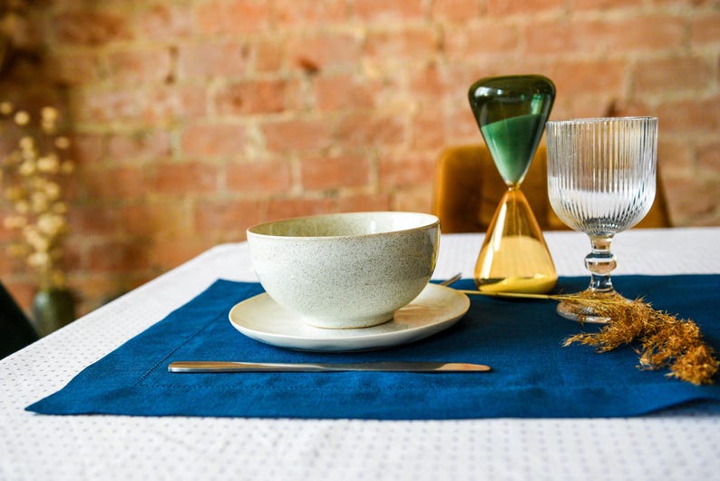 Puede incluir: Un cuenco de cer&aacute;mica blanca con un acabado moteado se encuentra sobre un mantel individual azul con un tenedor plateado y un reloj de arena dorado en el fondo. El mantel individual est&aacute; sobre un mantel blanco con un patr&oacute;n de lunares blancos y negros.
