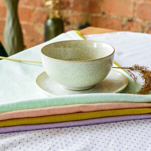 May include: A stack of five pastel-colored linen napkins on a white tablecloth with a speckled pattern. A speckled ceramic bowl sits on a white saucer on top of the napkins.