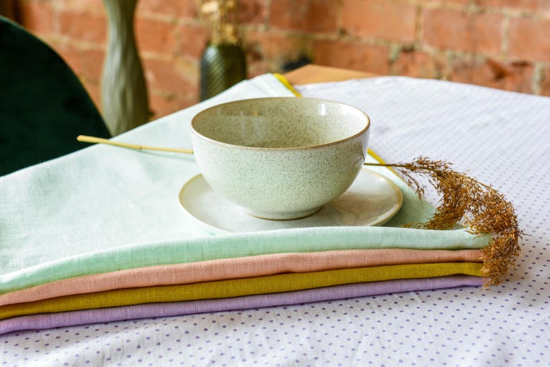 May include: A stack of five pastel-colored linen napkins on a white tablecloth with a speckled pattern. A speckled ceramic bowl sits on a white saucer on top of the napkins.