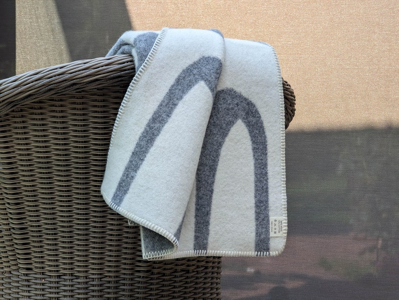 May include: A white and gray wool throw blanket with a geometric pattern of arches. The blanket is folded and draped over the back of a wicker chair.