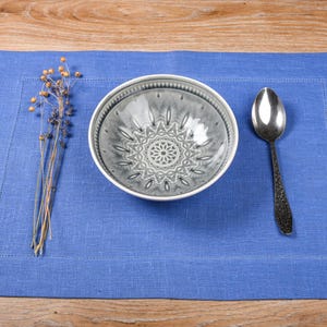 May include: A blue linen placemat with a white bowl with a gray patterned design and a silver spoon.