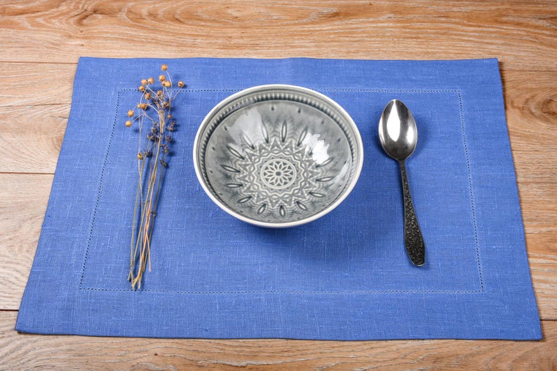 May include: A blue linen placemat with a white bowl with a gray patterned design and a silver spoon.
