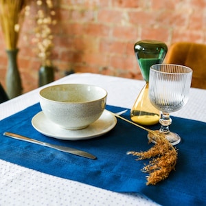 Puede incluir: Un mantel individual azul con un taz&oacute;n blanco, un platillo blanco, un cuchillo plateado y un vaso de agua. Un tallo de flor seca est&aacute; sobre la mesa junto al vaso.
