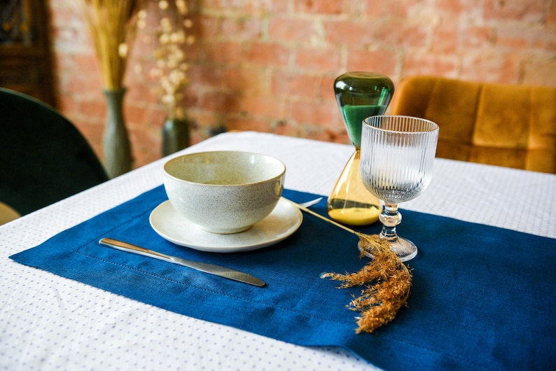 Puede incluir: Un mantel individual azul con un taz&oacute;n blanco, un platillo blanco, un cuchillo plateado y un vaso de agua. Un tallo de flor seca est&aacute; sobre la mesa junto al vaso.