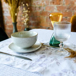 May include: A white ceramic bowl with a gray speckled glaze sits on a white saucer with a floral pattern. A clear glass with vertical ridges and a silver stem sits on a white tablecloth with a floral pattern. A brown and yellow dried flower arrangement is in the background.