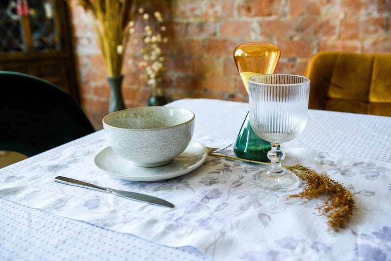 May include: A white ceramic bowl with a gray speckled glaze sits on a white saucer with a floral pattern. A clear glass with vertical ridges and a silver stem sits on a white tablecloth with a floral pattern. A brown and yellow dried flower arrangement is in the background.