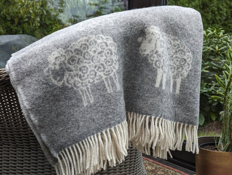 May include: A gray wool throw blanket with a white sheep design. The blanket has a fringe border.