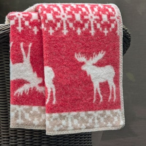 May include: Red and white patterned throw blanket with a moose and reindeer design. The blanket is folded and draped over a wicker chair.