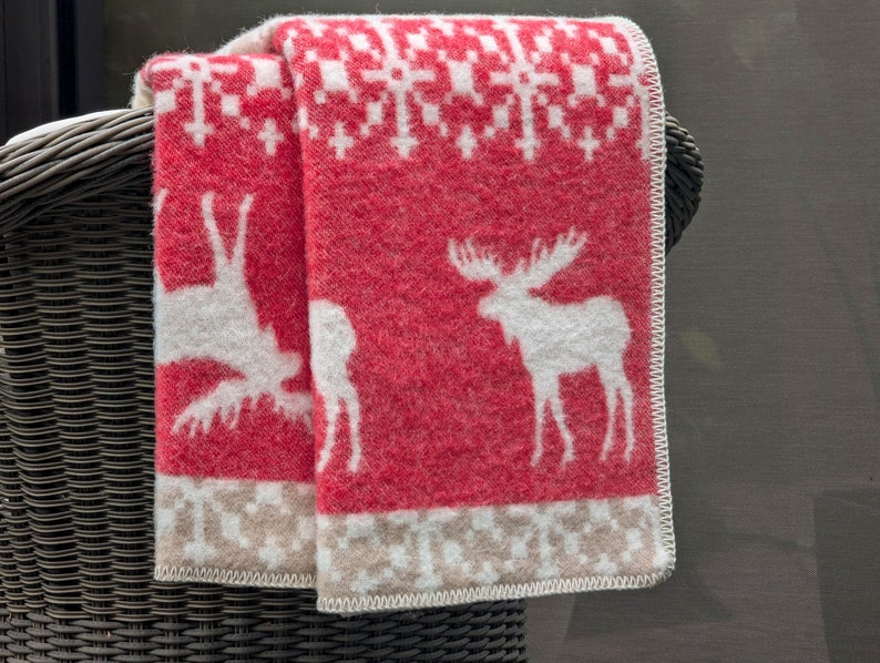 May include: Red and white patterned throw blanket with a moose and reindeer design. The blanket is folded and draped over a wicker chair.