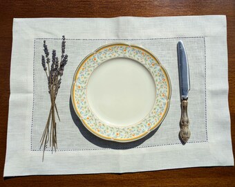 May include: A formal table setting featuring a white plate with a floral design, a silver knife, and a bundle of lavender on a linen placemat. The placemat has a stitched border and is set on a dark wooden surface.