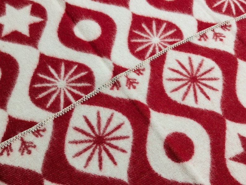 Puede incluir: Primer plano de una tela estampada en rojo y blanco. El dise&ntilde;o presenta formas de l&aacute;grima estilizadas, estrellas, c&iacute;rculos y elementos similares a copos de nieve. Una costura blanca en zigzag divide la tela diagonalmente.