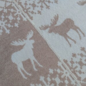 May include: A beige and white woven throw blanket with a repeating pattern of moose and snowflakes. The blanket has a fringed edge.