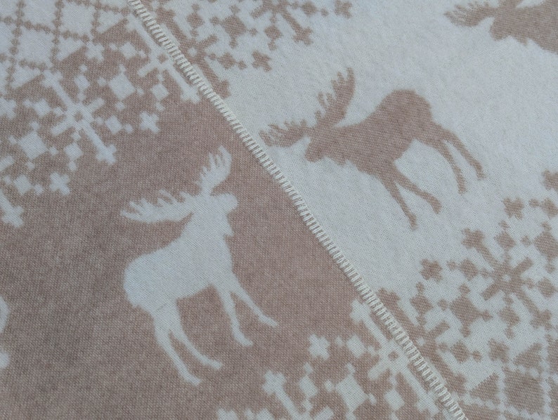 May include: A beige and white woven throw blanket with a repeating pattern of moose and snowflakes. The blanket has a fringed edge.
