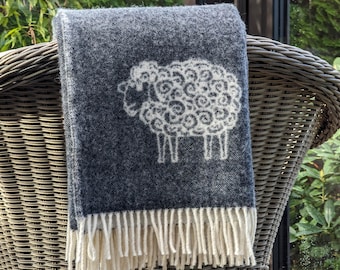 May include: A dark charcoal gray woven throw blanket featuring a white line-art illustration of a sheep with curly wool. The blanket has a textured appearance and is finished with a row of cream-colored fringe along the bottom edge.