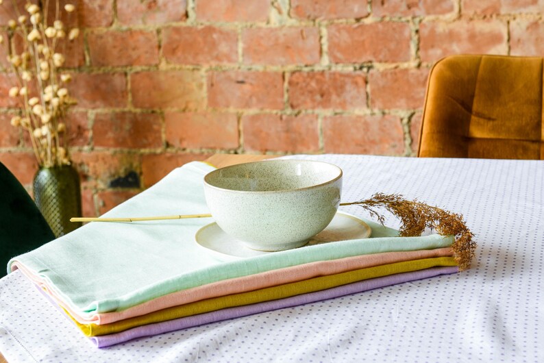 May include: A set of four pastel-colored linen napkins stacked on a white tablecloth with a small white bowl and a dried flower stem.