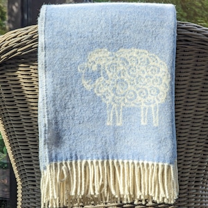 Può includere: Una coperta in lana blu chiaro con un design di pecora bianca e frange. La coperta è drappeggiata su una sedia in vimini.