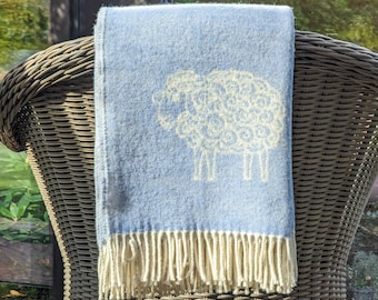 May include: A light blue throw blanket with a white sheep design and fringe trim. The blanket is draped over a wicker chair.