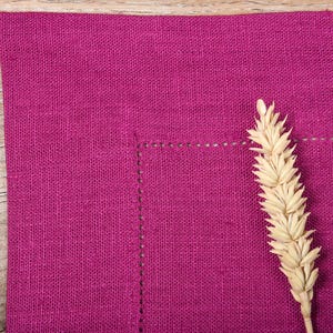 May include: A close-up of a deep pink linen placemat with a single wheat stalk in the center. The placemat has a subtle texture and a stitched border.