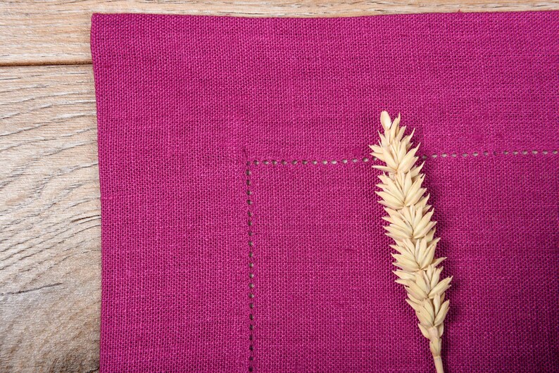 May include: A close-up of a deep pink linen placemat with a single wheat stalk in the center. The placemat has a subtle texture and a stitched border.