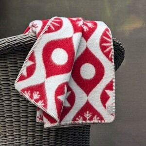 Puede incluir: Una manta plegada con estampado rojo y blanco, sobre una silla de mimbre. La manta presenta un dise&ntilde;o repetitivo de formas de l&aacute;grima rojas con c&iacute;rculos blancos y estrellas. Los bordes est&aacute;n acabados con costuras blancas.