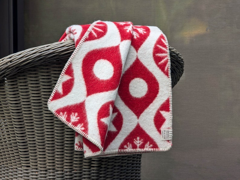 Puede incluir: Una manta plegada con estampado rojo y blanco, sobre una silla de mimbre. La manta presenta un dise&ntilde;o repetitivo de formas de l&aacute;grima rojas con c&iacute;rculos blancos y estrellas. Los bordes est&aacute;n acabados con costuras blancas.