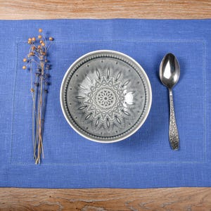 May include: A blue linen placemat with a gray ceramic bowl and a silver spoon. The bowl has a decorative pattern.
