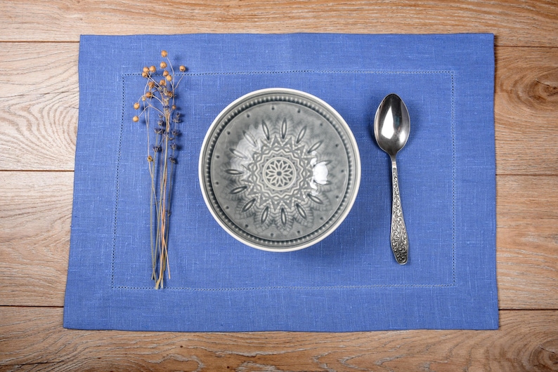 May include: A blue linen placemat with a gray ceramic bowl and a silver spoon. The bowl has a decorative pattern.