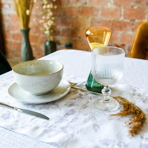 May include: A white tablecloth with a floral pattern, a gray speckled bowl with a white saucer, a clear glass with vertical ridges, and a yellow glass vase with a green base.