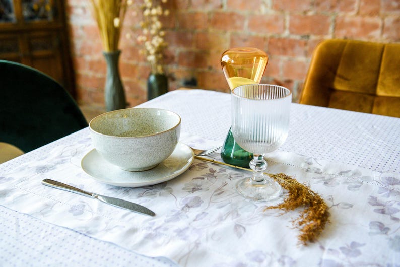 May include: A white tablecloth with a floral pattern, a gray speckled bowl with a white saucer, a clear glass with vertical ridges, and a yellow glass vase with a green base.