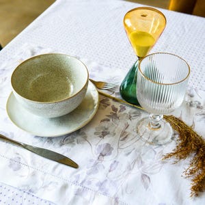 May include: A white tablecloth with a floral pattern, a gray ceramic bowl with a saucer, a glass of water, a silver knife, and a yellow hourglass with a green base.