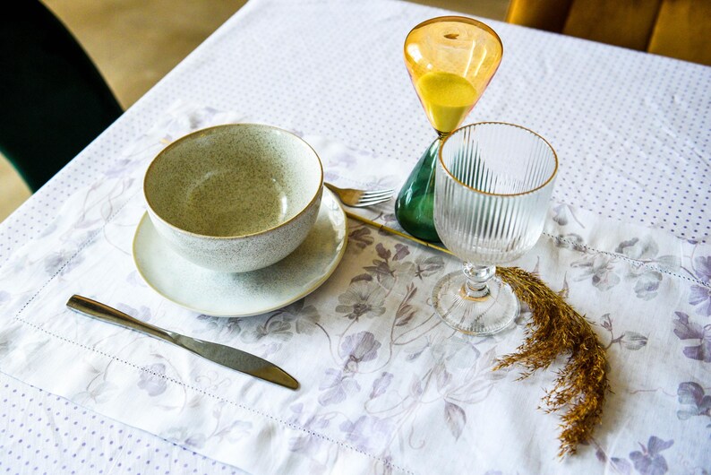 May include: A white tablecloth with a floral pattern, a gray ceramic bowl with a saucer, a glass of water, a silver knife, and a yellow hourglass with a green base.