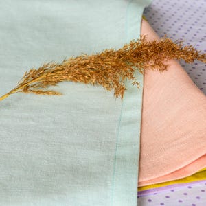 May include: A stack of four linen napkins in shades of light blue, peach, yellow, and purple. The napkins are folded and arranged on a purple background with white polka dots. A dried grass stalk is placed on top of the napkins.