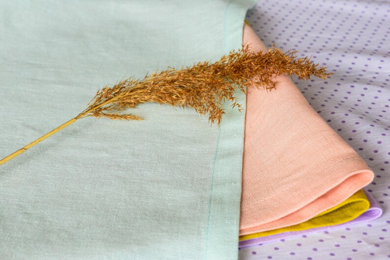 May include: A stack of four linen napkins in shades of light blue, peach, yellow, and purple. The napkins are folded and arranged on a purple background with white polka dots. A dried grass stalk is placed on top of the napkins.