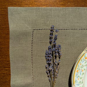 Puede incluir: Un mantel individual de lino de color marr&oacute;n claro con un borde decorativo cosido. Un ramo de flores de lavanda secas se coloca sobre el mantel individual, y un plato de porcelana blanco con un dise&ntilde;o floral es parcialmente visible en el fondo.