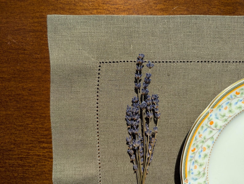 Puede incluir: Un mantel individual de lino de color marr&oacute;n claro con un borde decorativo cosido. Un ramo de flores de lavanda secas se coloca sobre el mantel individual, y un plato de porcelana blanco con un dise&ntilde;o floral es parcialmente visible en el fondo.