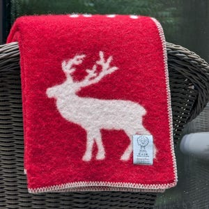 May include: A red wool throw blanket with a white reindeer design. The blanket has a white border and a small white tag with the text "Svensk Ull" and "Living & Giving" on it.