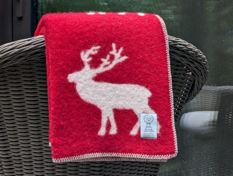 May include: A red wool throw blanket with a white reindeer design. The blanket has a white border and a small white tag with the text "Svensk Ull" and "Living & Giving" on it.