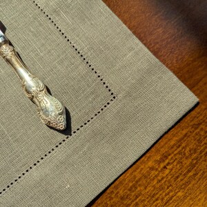 Puede incluir: Un cuchillo de mantequilla de plata con un mango adornado descansa sobre un mantel individual de lino beige. El mantel individual tiene un borde decorativo cosido.